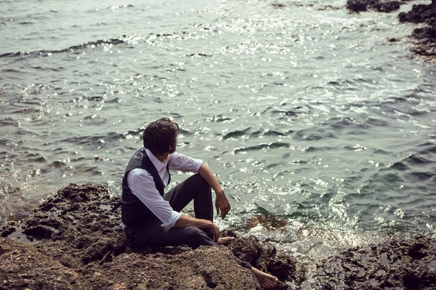 L'uomo con la barba in abiti scuri e una camicia bianca si siede sulla spiaggia di pietra in Crimea tarkhankut