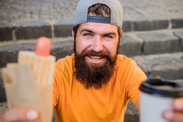 L'uomo con la barba mangia gustose salsicce e beve un bicchiere di carta Concetto di cibo di strada Stile di vita urbano Nutrizione Cibo spazzatura Gli hipster spensierati mangiano cibo spazzatura mentre si siedono le scale Spuntino di buon umore Ragazzo che mangia hot dog