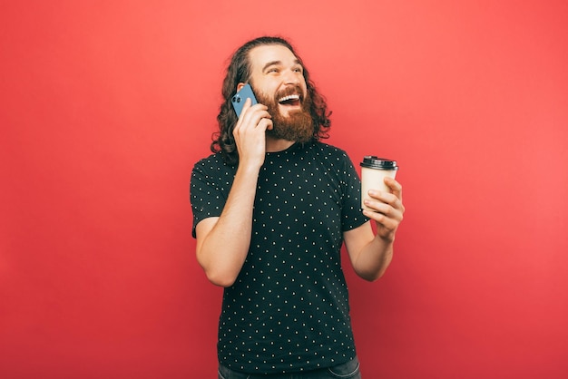 L'uomo con la barba tiene in mano il suo caffè e parla in modo eccitante al suo telefono su sfondo rosso