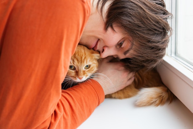 L'uomo con la maglietta arancione sta abbracciando un gatto rosso carino, un animale domestico adottivo, un ex gatto randagio a casa, un animale domestico soffice.