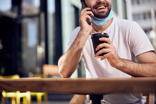 L'uomo con la maschera abbassata parla con lo smartphone e beve caffè