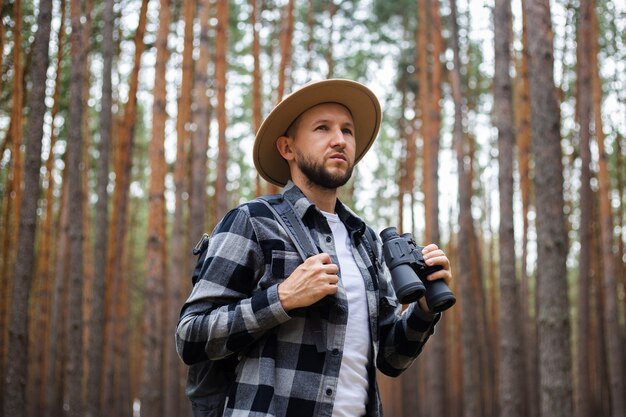 L'uomo con un cappello e una camicia a quadri tiene un binocolo e cammina attraverso la foresta.