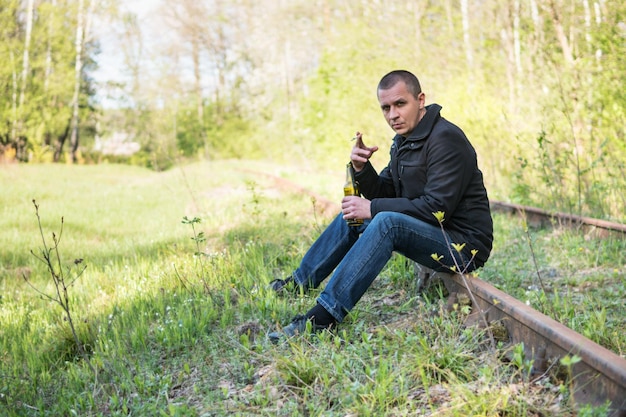 L'uomo corto si siede sui binari nella foresta con una sigaretta e una bottiglia in mano