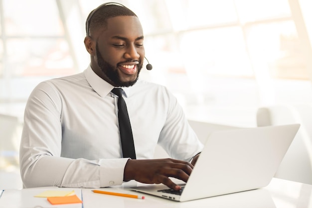 L'uomo d'affari afroamericano bello in vestito e auricolare sorride mentre lavora con il computer portatile in ufficio