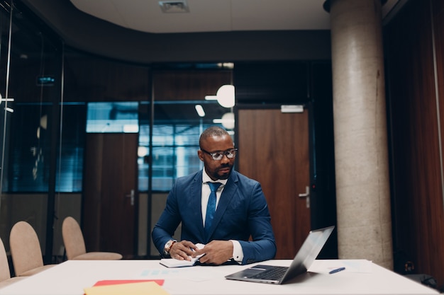 L'uomo d'affari afroamericano sorridente del ritratto in vestito blu si siede alla tavola per l'incontro in ufficio con il taccuino con la penna ed il computer portatile.