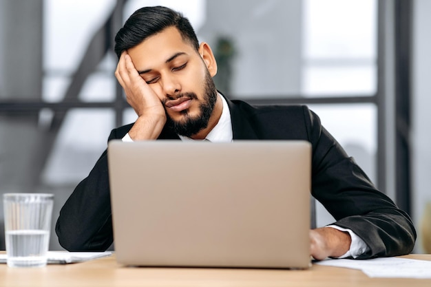 L'uomo d'affari arabo o indiano assonnato in giacca e cravatta si siede sul posto di lavoro in ufficio con un laptop si è messo la testa sulla mano si è addormentato durante il lavoro stanco oberato di lavoro esperienze cronica mancanza di sonno