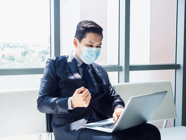 L'uomo d'affari asiatico in vestito che indossa la maschera protettiva ha alzato la mano felice e allegro quando ha visto un monitor portatile sulle sue ginocchia