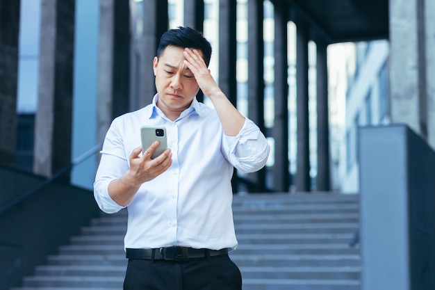 L'uomo d'affari asiatico sconvolto legge le notizie dall'uomo dello schermo del telefono deluso dalle cattive notizie