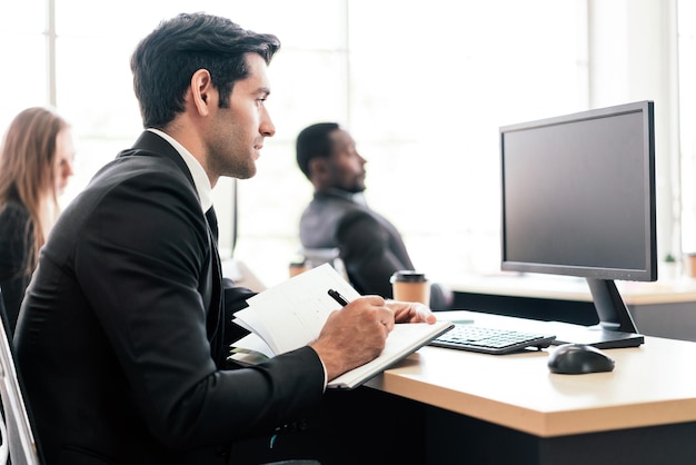 L'uomo d'affari caucasico scrive qualcosa su un piccolo quaderno mentre guarda il monitor del computer