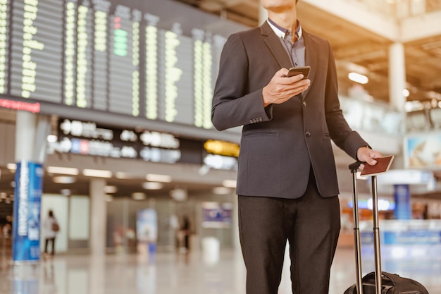 L'uomo d'affari che sta al tabellone per le affissioni di orario di volo tiene lo smartphone al cancello del terminal dell'aeroporto.