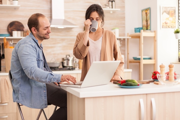 L'uomo d'affari che utilizza il computer portatile in cucina e wi fe sta bevendo una tazza di caffè. Felice amorevole allegro romantico innamorato coppia a casa utilizzando la moderna tecnologia internet wireless wifi