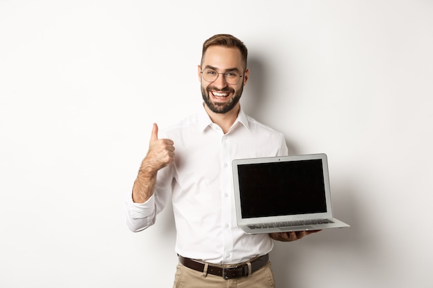 L'uomo d'affari di successo che mostra lo schermo del laptop, compone il pollice in approvazione, elogia qualcosa di buono, in piedi su sfondo bianco.