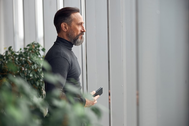 L'uomo d'affari è in piedi vicino a grandi finestre nello spazio ufficio e tiene lo smartphone
