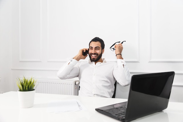 L'uomo d'affari felice con la barba bello indossa una camicia bianca tiene gli occhiali in mano, parla al telefono nell'interno dell'ufficio bianco