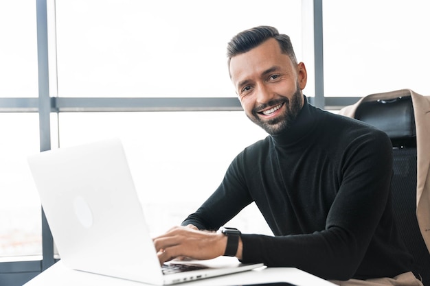 L'uomo d'affari lavora in ufficio dietro il laptop e guarda la telecamera con un sorriso