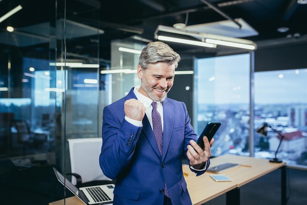 L'uomo d'affari senior ed esperto del leader soddisfatto e felice guarda il telefono sorride e si rallegra per la vittoria ricevuta una buona notizia