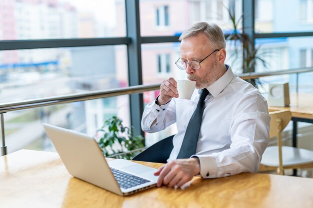 L'uomo d'affari senior indossa una camicia bianca e beve caffè vicino alla grande finestra con vista sulla città. Utilizzo del computer portatile.