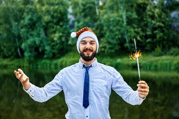 L'uomo d'affari sorridente di Houng, che indossa un cappello di Babbo Natale, tiene in mano una scintilla ardente, in piedi sullo sfondo di alberi verdi e fiume in natura.