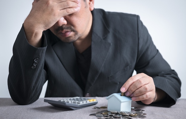 L'uomo d'affari sta mettendo le monete nel salvadanaio di una piccola casa e si sente stressato quando sa che non ha abbastanza soldi per pagare le rate della casa