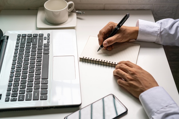 L'uomo d'affari sta prendendo nota sul taccuino dall'imformazione finanziaria dal computer portatile Concetto del lavoro di affari.