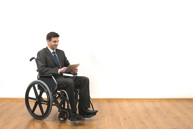 L'uomo d'affari sulla sedia a rotelle che tiene una compressa su un fondo bianco della parete