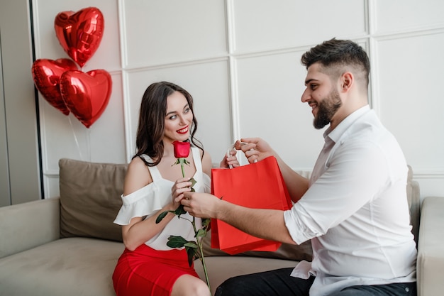 L'uomo dà borse della spesa e rosa rossa alla donna a casa con palloncini a forma di cuore sul divano