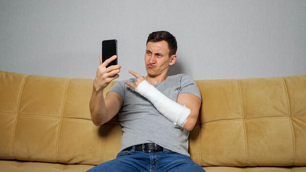 L'uomo dai capelli castani fa selfie e posa con l'osso del raggio ferito in una benda di gesso utilizzando lo smartphone seduto sul divano in soggiorno