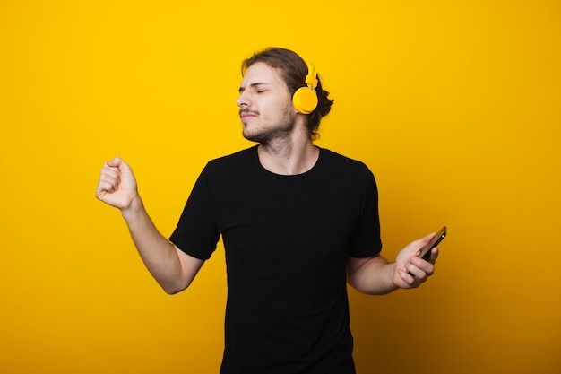 L'uomo dai capelli lunghi con la barba sta ballando indossando le cuffie e tenendo un cellulare su una parete gialla di uno studio che indossa una maglietta nera