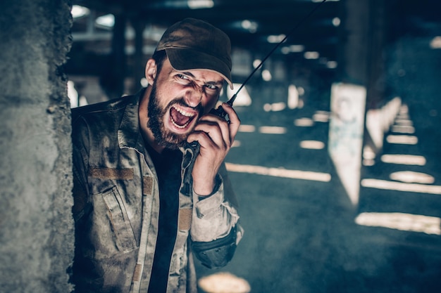 L'uomo dall'esercito tiene la radio portatile vicino al viso e parla a voce molto alta. Sta urlando. Guy è seduto dietro la colonna e guarda dritto in avanti. Si sta nascondendo.