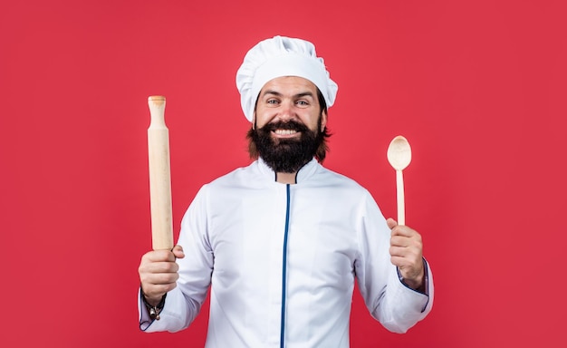 L'uomo del cuoco barbuto del ristorante professionale tiene il mattarello di legno e l'utensile da cucina del cucchiaio