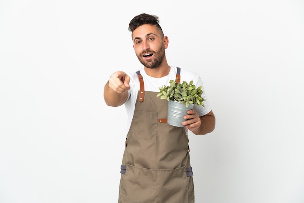 L'uomo del giardiniere che tiene una pianta sopra la parete bianca isolata ha sorpreso e che indica la parte anteriore