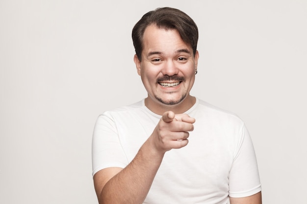 L'uomo della felicità, che indossa una maglietta bianca, ha un aspetto felice, punta il dito verso la telecamera e sorride a trentadue denti. Studio isolato girato su sfondo grigio