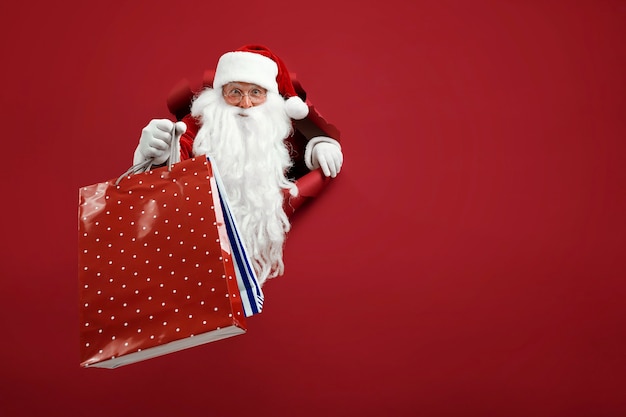 L'uomo della Santa tiene in mano il pacchetto dei negozi attraverso un buco della carta. Uomo barbuto in santa hat guardando attraverso il foro su carta rossa.