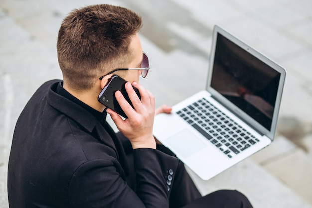 L'uomo di affari si siede sulle scale fuori dell'ufficio, lavorando al computer portatile, chiamando il telefono. Elegante modello maschile in giacca nera. Giovane con un telefono cellulare che si siede sui gradini