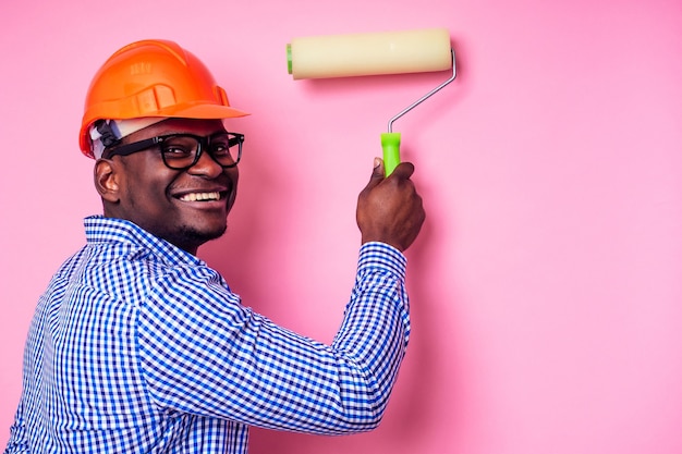 L'uomo di colore afroamericano che tiene il rullo di vernice in mano dipinge il muro di colore rosa. Felice costruttore africano che dipinge all'interno della casa, l'uomo d'affari indossa un casco duro. Il giovane ragazzo sta dipingendo