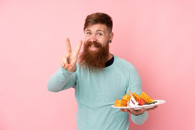 L'uomo di redhead con la barba lunga che tiene i waffles sopra la parete dentellare isolata che sorride e che mostra il segno di vittoria