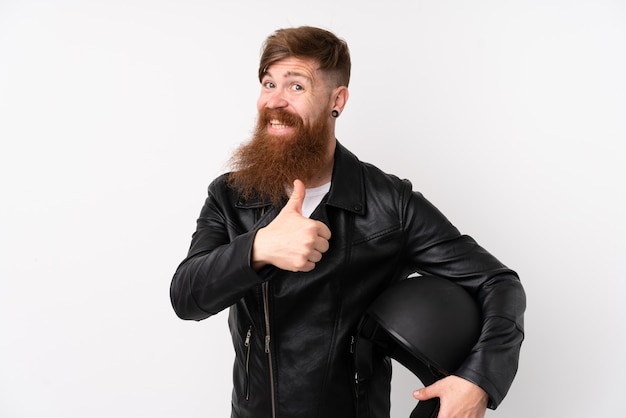 L'uomo di Redhead con la barba lunga che tiene un casco del motociclo sopra la parete bianca isolata che dà un pollice aumenta il gesto