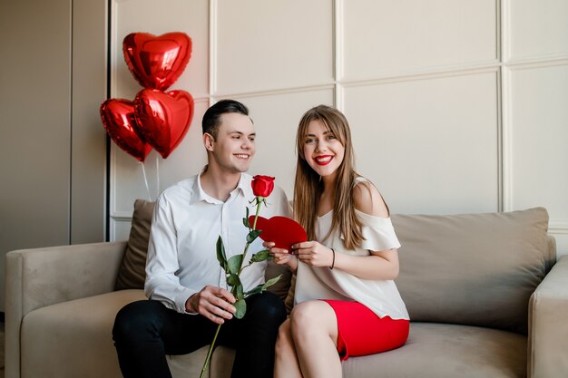 L'uomo e la donna a casa sul divano con la rosa rossa e il cuore hanno modellato la carta del biglietto di S. Valentino