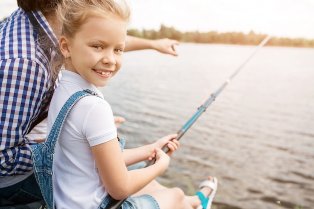 L'uomo è seduto sulla riva del fiume con sua figlia e punta in avanti. La ragazza sta tenendo l'asta di pesce e sta esaminando la macchina fotografica. Lei sta sorridendo. La ragazza sembra felice.