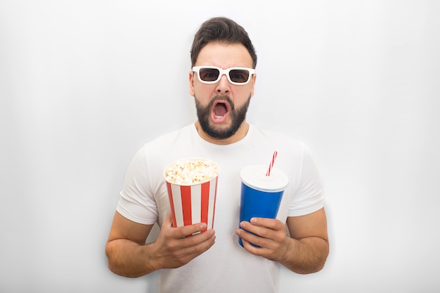 L'uomo eccitato tiene la bocca aperta. Tiene contemporaneamente un secchio di popcorn e un bicchiere di cola. Guy indossa occhiali da film.