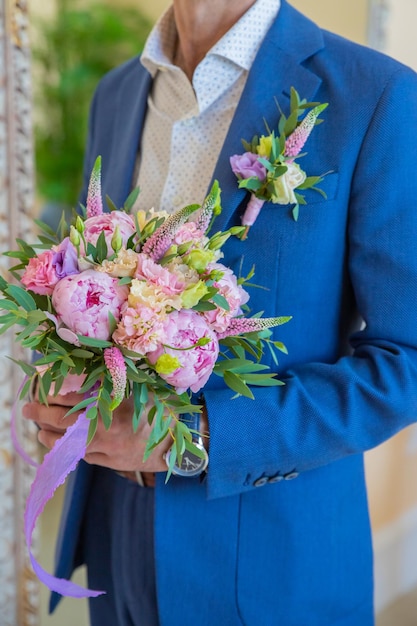 L'uomo elegante in abito tiene un bouquet rotondo con peonie ed eustomaBoutonniere di eremurus eustoma bianco e verde attaccati alla giacca blu dello sposo Dettagli del matrimonio e floristica