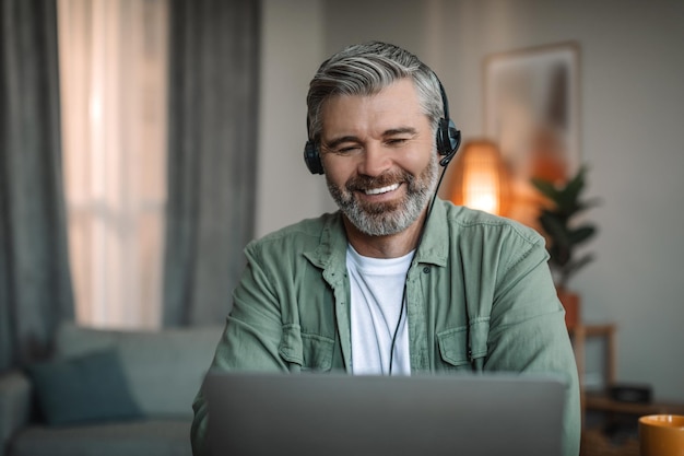 L'uomo europeo anziano sorridente con la barba in cuffia guarda il discorso del laptop con la lezione video del cliente