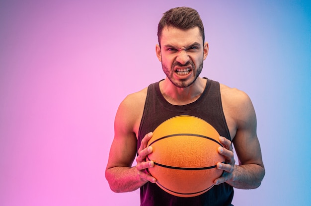 L'uomo europeo arrabbiato tiene la palla da basket nelle mani. Il giovane ragazzo barbuto indossa la canottiera e guarda la telecamera. Isolato su sfondo blu e rosa. Riprese in studio. Copia spazio