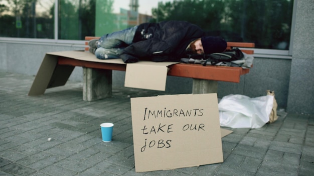 L'uomo europeo senza casa e senza lavoro con cartello di cartone dorme su una panchina in una strada di città