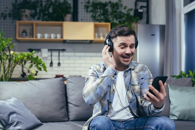 L'uomo felice a casa che sorride e ascolta la musica dal telefono utilizza l'app musicale sullo smartphone online con grandi cuffie