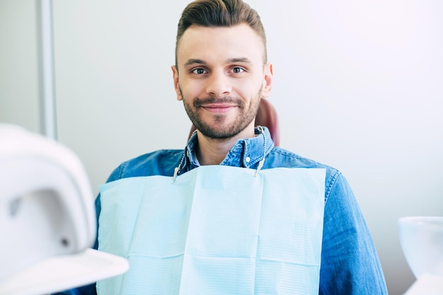 L'uomo felice con gli occhi nocciola e i capelli castano scuro è seduto sulla poltrona del dentista e sorride dritto nella telecamera perché è soddisfatto del lavoro di un dentista