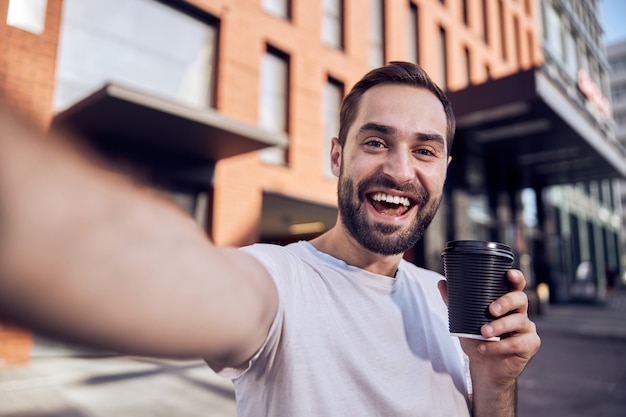 L'uomo felice con il caffè prende il selfie all'aperto