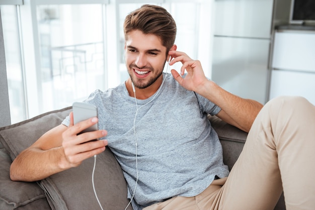 L'uomo felice in auricolari ascolta la musica mentre si trova sul divano. Guardando il telefono e la messaggistica.