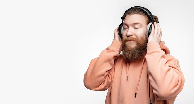 L'uomo felice scandinavo ha chiuso gli occhi e ascolta la musica in cuffie professionali isolate su sfondo grigio Ragazzo felice con barba e acconciatura allo zenzero Tecnologie digitali moderne