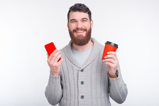 L'uomo felice sta tenendo il suo telefono e una tazza rossa per andare su fondo bianco.